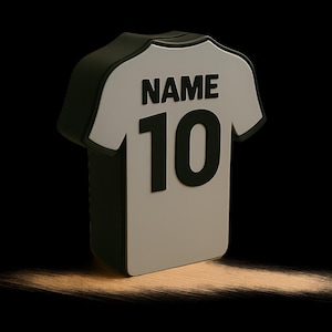 K&ouml;nnte beinhalten: 3D-Darstellung eines wei&szlig;en Fu&szlig;balltrikots mit der Nummer 10 und dem Wort "NAME" in Schwarz auf dem R&uuml;cken. Das Trikot hat schwarze Details. Der Hintergrund ist schwarz mit einer hellbraunen, strukturierten Oberfl&auml;che.