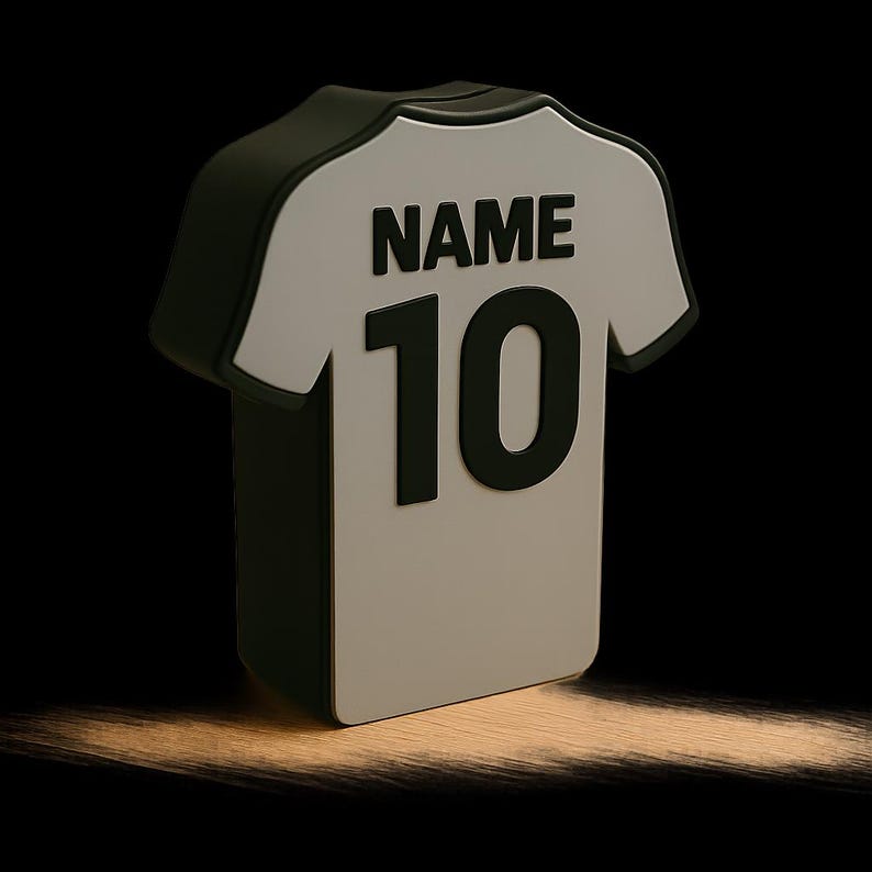 K&ouml;nnte beinhalten: 3D-Darstellung eines wei&szlig;en Fu&szlig;balltrikots mit der Nummer 10 und dem Wort "NAME" in Schwarz auf dem R&uuml;cken. Das Trikot hat schwarze Details. Der Hintergrund ist schwarz mit einer hellbraunen, strukturierten Oberfl&auml;che.
