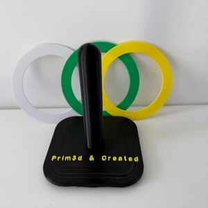 May include: A black 3D-printed paper towel holder with a vertical post and a square base. The base has the text "Prim3d & Created" in yellow. Behind the holder are three colored rings: white, green, and yellow.