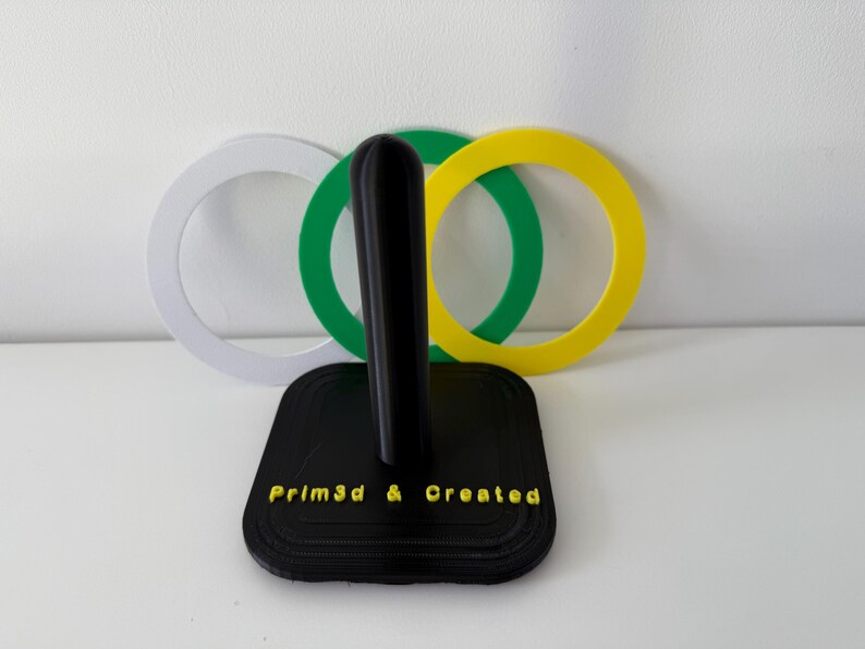 May include: A black 3D-printed paper towel holder with a vertical post and a square base. The base has the text "Prim3d & Created" in yellow. Behind the holder are three colored rings: white, green, and yellow.