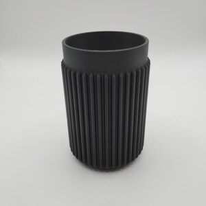 May include: A black, cylindrical tumbler with a ribbed exterior and a smooth rim. The cup is made of a matte material and is designed to hold beverages. The design is simple and modern, suitable for everyday use.