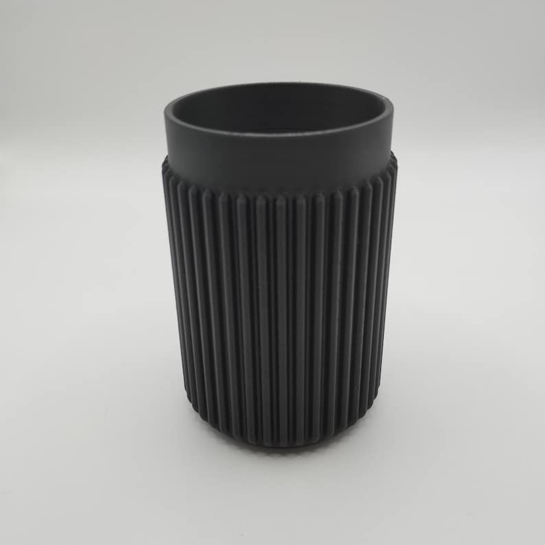 May include: A black, cylindrical tumbler with a ribbed exterior and a smooth rim. The cup is made of a matte material and is designed to hold beverages. The design is simple and modern, suitable for everyday use.