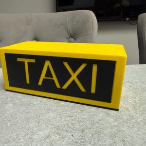 May include: A yellow taxi cab roof sign with the word "TAXI" in bold, yellow letters on a black background. The sign is rectangular and sits on a gray surface.