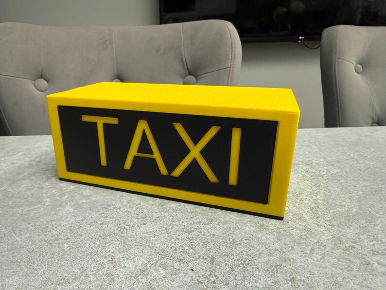 May include: A yellow taxi cab roof sign with the word "TAXI" in bold, yellow letters on a black background. The sign is rectangular and sits on a gray surface.