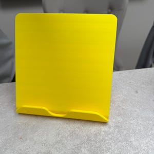 May include: A bright yellow tablet or book stand. The stand is made of a solid material and has a rectangular shape with rounded corners. It features a small lip at the bottom to hold a tablet or book in place.