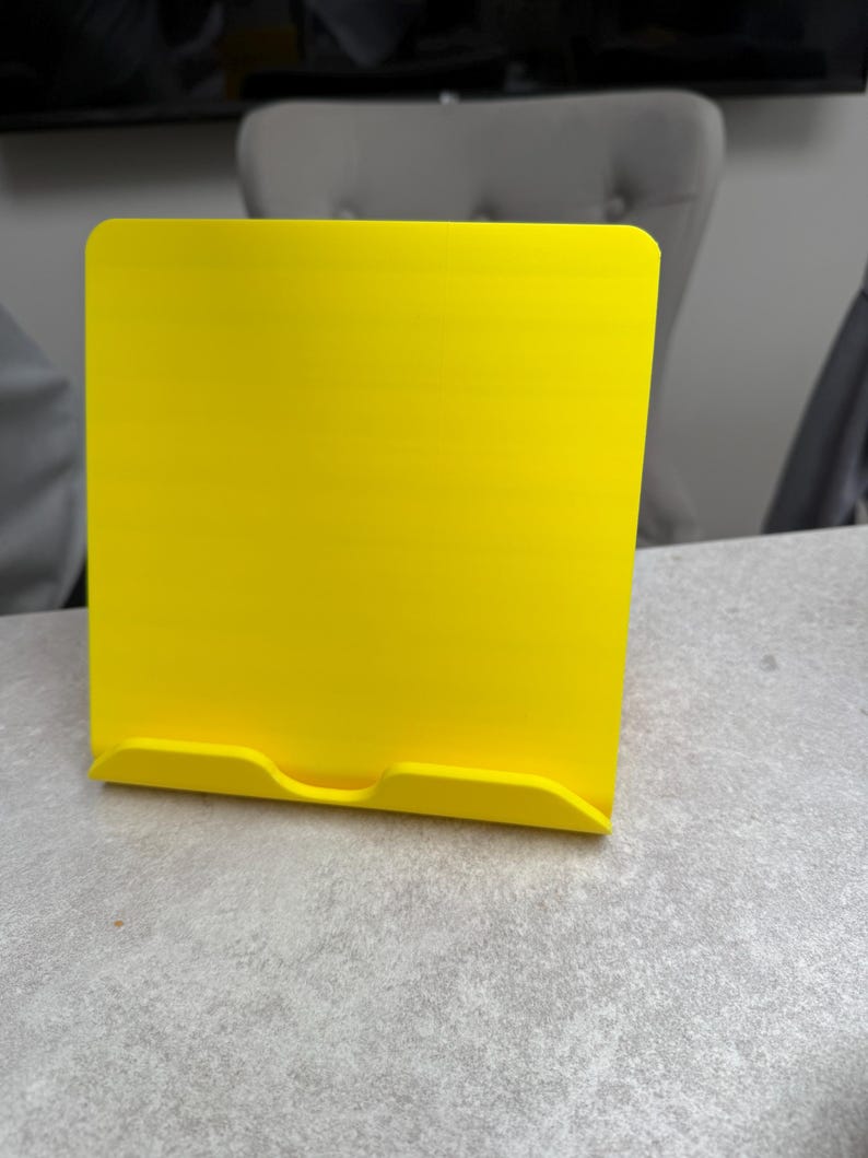 May include: A bright yellow tablet or book stand. The stand is made of a solid material and has a rectangular shape with rounded corners. It features a small lip at the bottom to hold a tablet or book in place.