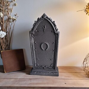 May include: A dark gray tombstone-shaped decoration with an arched top and a decorative base. The tombstone features a central circular design and star and line accents. The item is displayed on a wooden surface.