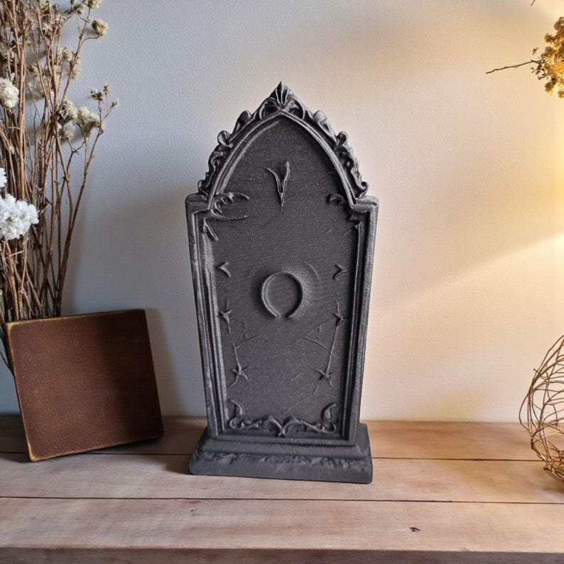 May include: A dark gray tombstone-shaped decoration with an arched top and a decorative base. The tombstone features a central circular design and star and line accents. The item is displayed on a wooden surface.