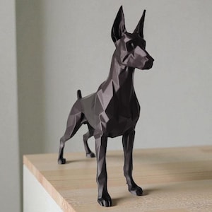 Op de afbeelding: Een donkerbruine geometrische sculptuur van een Dobermann Pinscher hond. De hond staat op een licht houten oppervlak, met een witte kastbasis. De sculptuur heeft een modern, hoekig ontwerp, met puntige oren en een strakke, minimalistische esthetiek.