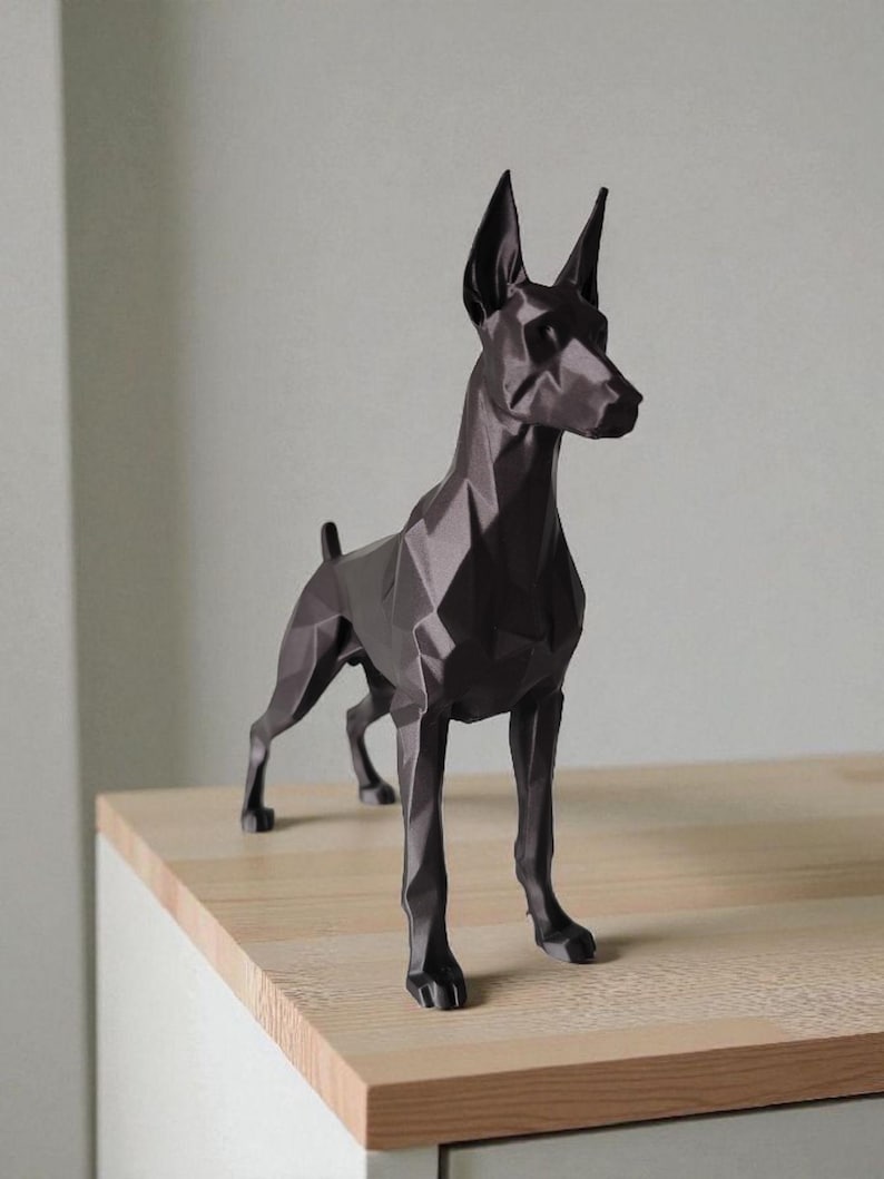 Op de afbeelding: Een donkerbruine geometrische sculptuur van een Dobermann Pinscher hond. De hond staat op een licht houten oppervlak, met een witte kastbasis. De sculptuur heeft een modern, hoekig ontwerp, met puntige oren en een strakke, minimalistische esthetiek.