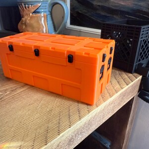 May include: Bright orange storage container with black latches and handles. The rectangular box sits on a wooden surface. A black plastic crate with legs is next to the container.