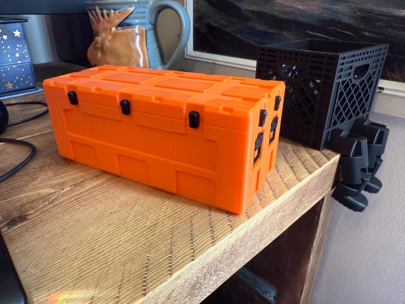 May include: Bright orange storage container with black latches and handles. The rectangular box sits on a wooden surface. A black plastic crate with legs is next to the container.