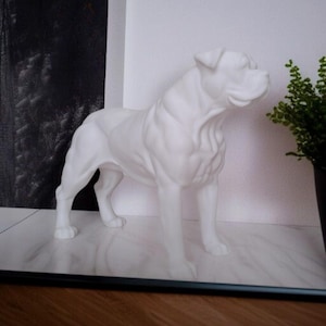 May include: A white ceramic dog statue in a standing pose. The sculpture has a smooth finish and detailed musculature, with the dog's head turned slightly to the side. The statue is placed on a marble-patterned surface.