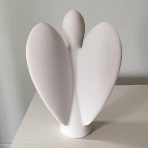 May include: A white ceramic angel figurine with outstretched wings. The sculpture has a smooth, matte finish and a minimalist design. The angel is standing on a round base, and the wings are curved and symmetrical.