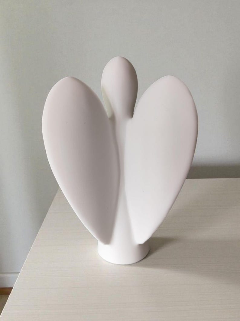 May include: A white ceramic angel figurine with outstretched wings. The sculpture has a smooth, matte finish and a minimalist design. The angel is standing on a round base, and the wings are curved and symmetrical.