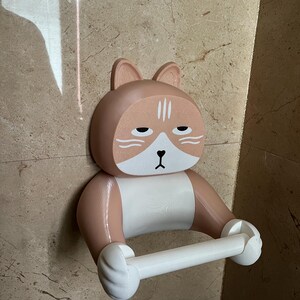 May include: A novelty cat-shaped toilet paper holder in shades of tan, white, and black. The holder is mounted on a wall and features a cat face with a sleepy expression. The toilet paper roll is held between the cat's paws.