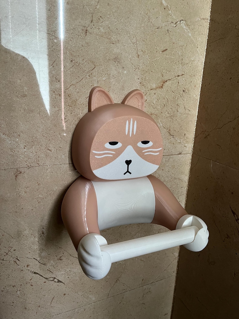 May include: A novelty cat-shaped toilet paper holder in shades of tan, white, and black. The holder is mounted on a wall and features a cat face with a sleepy expression. The toilet paper roll is held between the cat's paws.