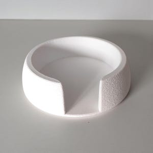 May include: A white, circular coaster with a textured exterior and a smooth interior. The coaster has a curved design with two straight cutouts on opposite sides. The object is made of a solid, opaque material.
