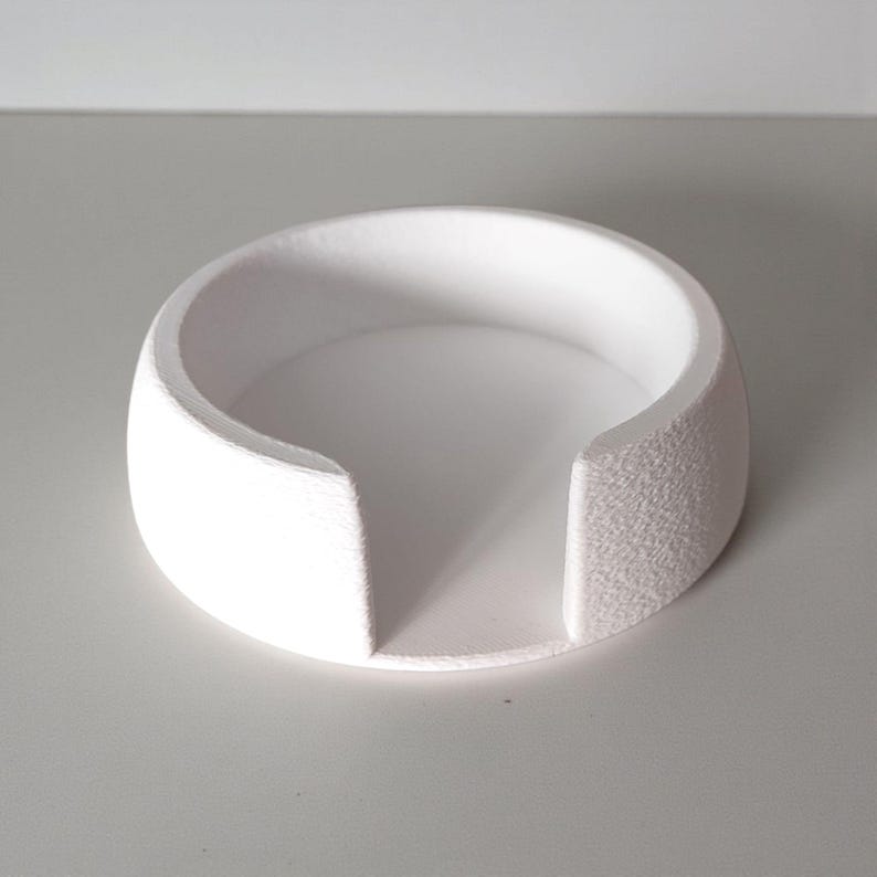 May include: A white, circular coaster with a textured exterior and a smooth interior. The coaster has a curved design with two straight cutouts on opposite sides. The object is made of a solid, opaque material.