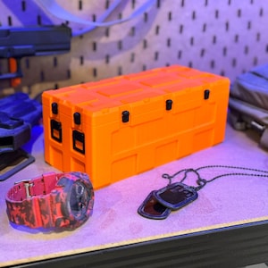May include: An orange storage case with black latches and hinges. A red and black camouflage watch and a black dog tag necklace are next to the case. The items are on a light purple surface.