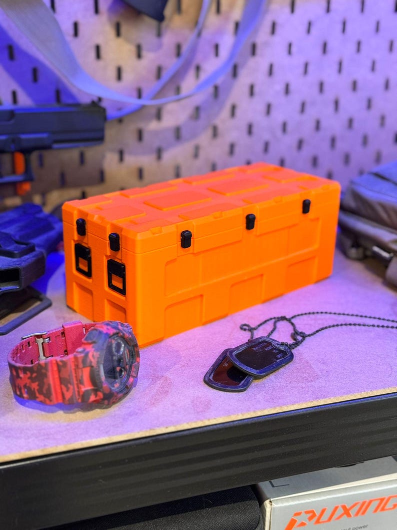 May include: An orange storage case with black latches and hinges. A red and black camouflage watch and a black dog tag necklace are next to the case. The items are on a light purple surface.