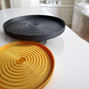 May include: Two decorative trays. The top tray is black and circular, with a raised, concentric ripple design. The bottom tray is gold-coloured, also circular, with a similar ripple pattern. Both trays are on a white surface.