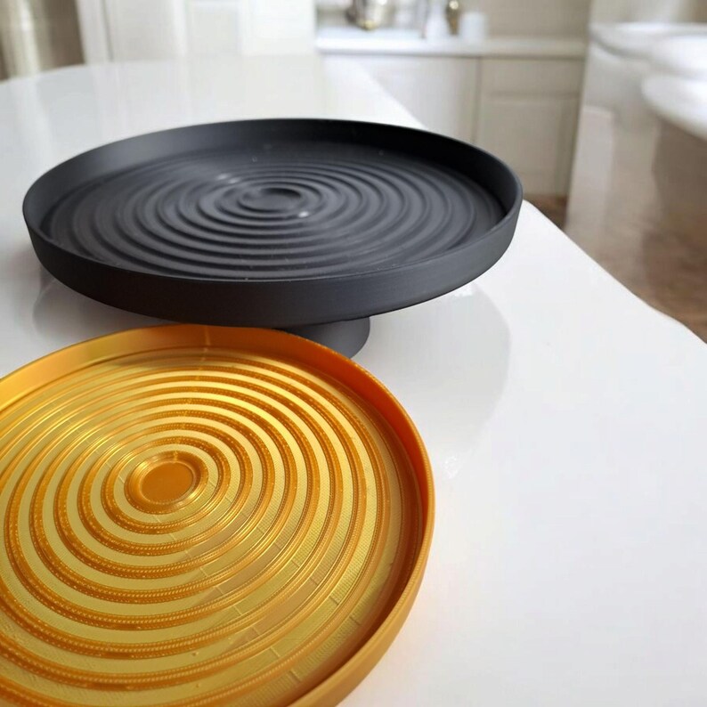 May include: Two decorative trays. The top tray is black and circular, with a raised, concentric ripple design. The bottom tray is gold-coloured, also circular, with a similar ripple pattern. Both trays are on a white surface.