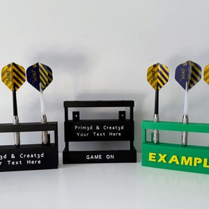 May include: Three dart holders in black and green, each holding three darts. The darts have yellow and blue flights. The black holders have the text "Prim3d & Creat3d Your Text Here" and the green holder has the text "EXAMPLE".