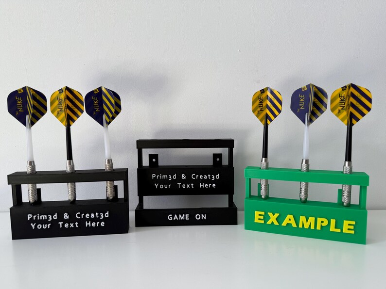May include: Three dart holders in black and green, each holding three darts. The darts have yellow and blue flights. The black holders have the text "Prim3d & Creat3d Your Text Here" and the green holder has the text "EXAMPLE".
