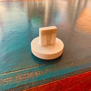 May include: A light-colored bottle stopper with a rectangular top and a circular base. The stopper is made of a speckled material and sits on a teal-colored table with a decorative border.