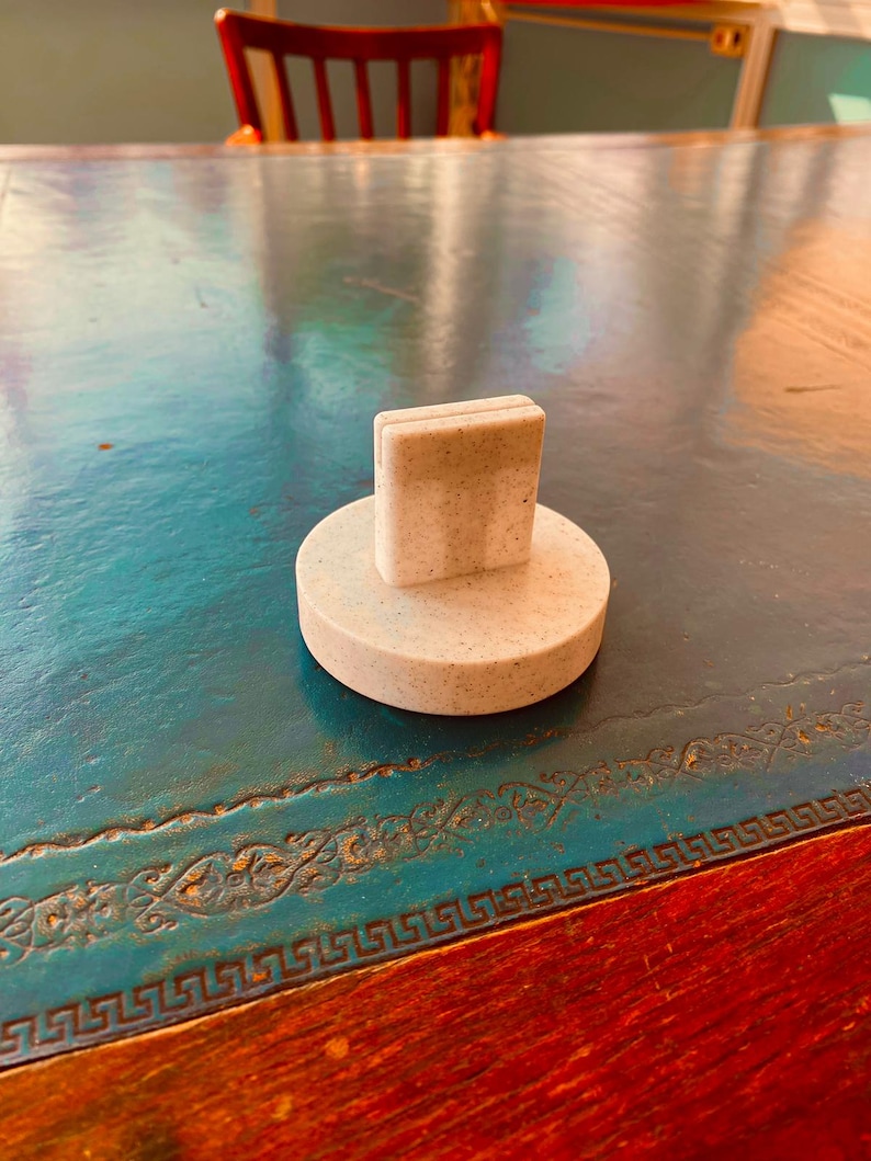 May include: A light-colored bottle stopper with a rectangular top and a circular base. The stopper is made of a speckled material and sits on a teal-colored table with a decorative border.