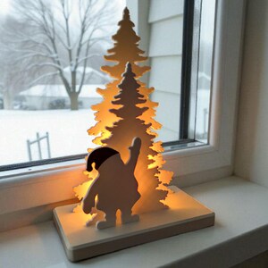 May include: A wooden Christmas decoration featuring a silhouette of Santa Claus with a raised arm, standing in front of a lit-up Christmas tree. The decoration is on a rectangular base and sits on a windowsill with a snowy outdoor scene visible.