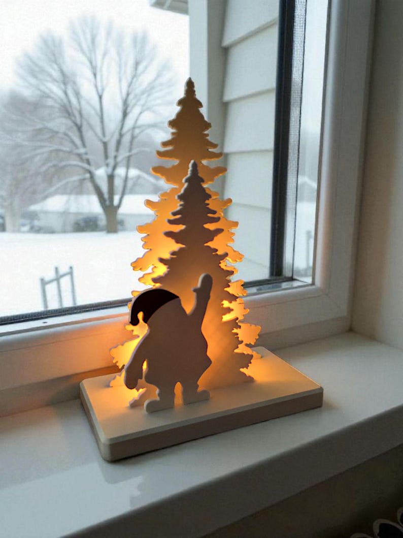 May include: A wooden Christmas decoration featuring a silhouette of Santa Claus with a raised arm, standing in front of a lit-up Christmas tree. The decoration is on a rectangular base and sits on a windowsill with a snowy outdoor scene visible.