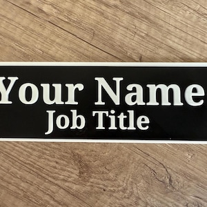 May include: A rectangular black and white nameplate. The nameplate has a black background with white text that reads "Your Name" on the top line and "Job Title" on the bottom line. The nameplate is framed with a white border.