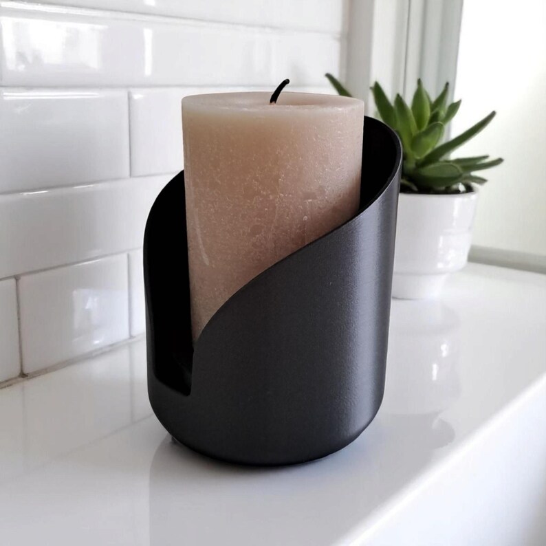 May include: A black metal candle holder with a beige pillar candle. The candle holder has a curved design that cradles the candle. A small succulent plant in a white pot is in the background. The candle holder is on a white surface.