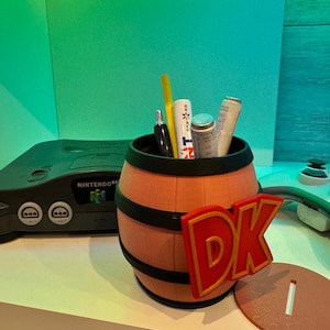 May include: A barrel-shaped pencil holder with the letters "DK" in red and gold. The pencil holder is filled with pens and other items. A Nintendo 64 console is in the background.