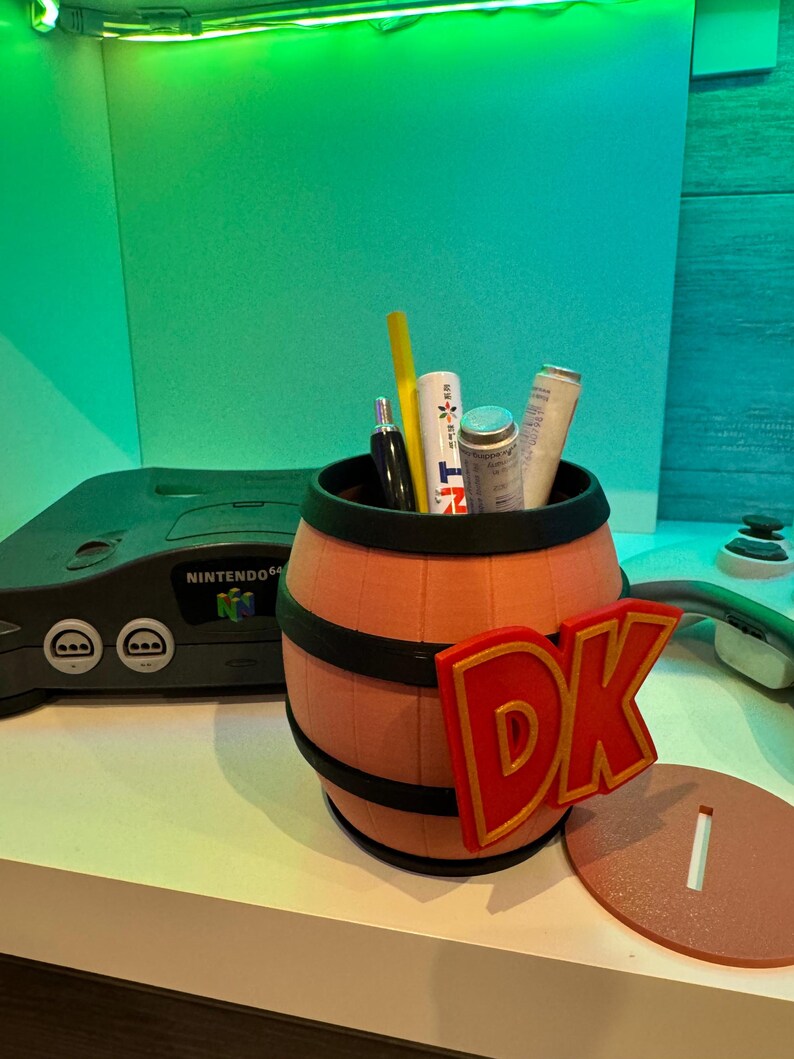 May include: A barrel-shaped pencil holder with the letters "DK" in red and gold. The pencil holder is filled with pens and other items. A Nintendo 64 console is in the background.