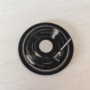 May include: A black and white round coaster featuring a vinyl record design. The coaster has a central white circle and a white needle arm. The design is printed on a black background.