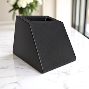May include: A matte black, geometric desk organizer with a rectangular opening at the top. The organizer has a slanted front and a flat base. The surface has a textured appearance. The background features a marble-like surface and a glimpse of white flowers.