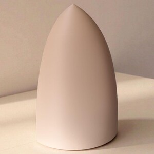 May include: A pale pink, teardrop-shaped object with a smooth, matte finish. The object has a rounded base and tapers to a sharp point at the top. It is set against a neutral background, highlighting its form and colour.