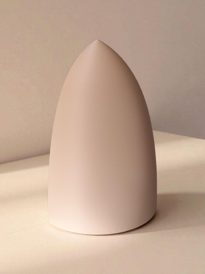 May include: A pale pink, teardrop-shaped object with a smooth, matte finish. The object has a rounded base and tapers to a sharp point at the top. It is set against a neutral background, highlighting its form and colour.