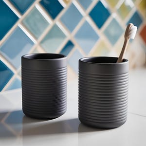 Pode incluir: Dois copos de casa de banho em cer&acirc;mica cinza escuro com detalhes de ranhuras horizontais. Um copo cont&eacute;m uma escova de dentes de madeira com cerdas brancas. Os copos est&atilde;o sobre uma superf&iacute;cie branca, com um fundo de azulejos coloridos.