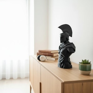 May include: A black geometric bust of a Spartan warrior wearing a helmet, placed on a light wood cabinet. Beside the bust are books, headphones, and a small potted plant. The bust is a decorative item.