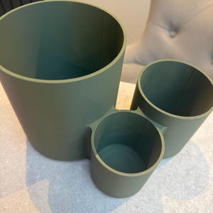 May include: A sage green desk organizer with three cylindrical compartments of varying sizes. The largest is on the left, with a medium and small cylinder attached. The organizer is made of a matte material.