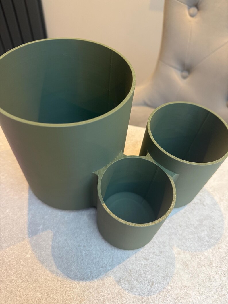 May include: A sage green desk organizer with three cylindrical compartments of varying sizes. The largest is on the left, with a medium and small cylinder attached. The organizer is made of a matte material.