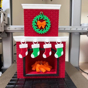 May include: A red brick-style Christmas fireplace decoration with a green wreath and orange bow. Four miniature stockings hang above the opening. Inside the fireplace are orange logs. The base is black.