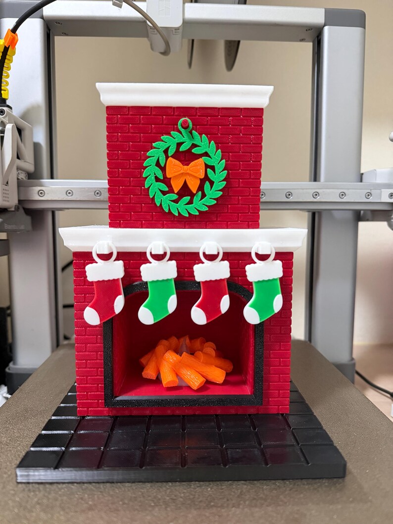 May include: A red brick-style Christmas fireplace decoration with a green wreath and orange bow. Four miniature stockings hang above the opening. Inside the fireplace are orange logs. The base is black.