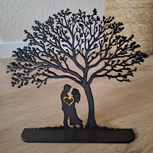 May include: Black metal tree silhouette with a couple embracing under its branches. A gold heart is centered between the figures. The tree is on a black base, suitable for display.