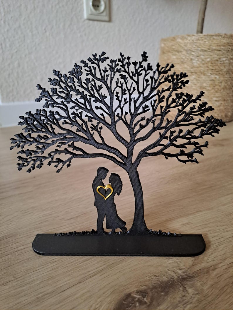 May include: Black metal tree silhouette with a couple embracing under its branches. A gold heart is centered between the figures. The tree is on a black base, suitable for display.