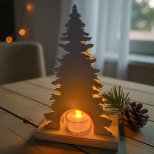 May include: A white wooden Christmas tree-shaped candle holder with a lit tea light inside. The candle holder is on a wooden table, next to a pine cone and a sprig of greenery. The background is blurred.